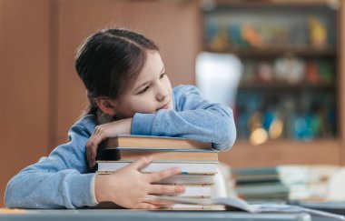 Parlak gözlü öğrenci derslerine kafa yoruyor, çenesini canlı bir sınıftaki kitap yığınının üzerinde dinlendiriyor. Atmosfer rahat, öğrenme ve keşfetme heyecanıyla dolu..