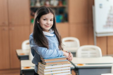 Genç bir öğrenci canlı bir sınıfta gururla duruyor, bir yığın kitabı dengeliyor. Onun neşeli ifadesi, öğrenme sevincini yaşarken okula duyduğu heyecanı yansıtıyor..
