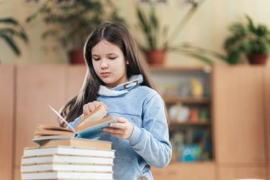 Bitkilerle dolu canlı bir sınıfta, bir kız öğrenci derinden bir kitaba odaklanıyor, sayfaları düşünceli bir şekilde çeviriyor. Yakınlardaki ders kitapları onun öğrenmeye olan bağlılığını simgeliyor..