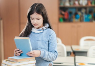 Genç bir öğrenci renkli bir sınıfta duruyor, dikkatle onun yanındaki kitaplara bakıyor. Atmosfer hayat dolu, öğrenme ve keşfetme heyecanıyla dolu..