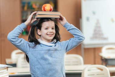 Neşeli bir liseli kız neşeli yanını kitaplarını dengeleyerek ve parlak, canlı bir sınıfta kafasında parlak bir elmayla gösterir. Öğrenmenin ve arkadaşlığın sevincini kutlar..