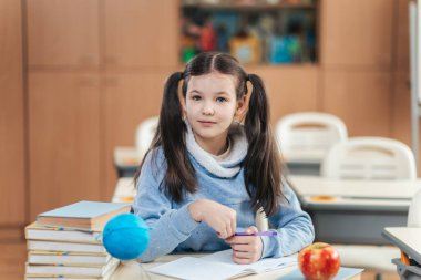 Parlak ve davetkar bir sınıfta örgülü saçlı bir kız öğrenci masasında oturup öğrenmeye hazır bir şekilde bekliyor. Etrafı kitap ve elmayla çevrili, bir kalem tutuyor ve kendinden emin bir şekilde ileri bakıyor..