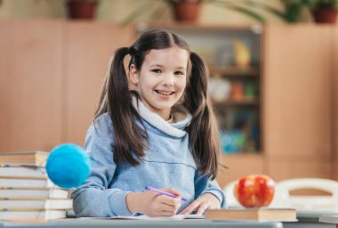 Saçları örgülü neşeli bir kız öğrenci masasında oturmuş, mutlu bir şekilde deftere yazıyor..