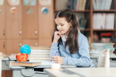 Canlı bir okul ortamında, odaklanmış bir öğrenci masasında oturur, yazarken derin düşüncelere dalardı. Etrafındaki okul malzemeleri öğrenme atmosferini canlandırıyor..