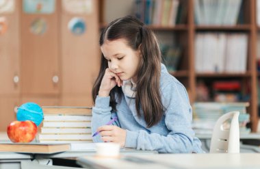 Canlı bir sınıfta, uzun saçlı genç bir kız öğrenci yazarken derin düşünür. Masasında oturuyor, etrafı kitaplarla ve renkli öğrenme araçlarıyla çevrili, çalışmalarına dalmış..