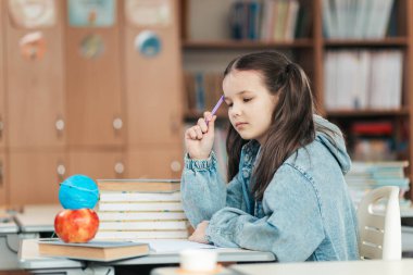Bir liseli kız, elinde kalemle masasında oturmuş renkli nesnelere bakarken derin düşüncelere dalıyor. Kitaplar ve parlak bir sınıf etrafını sarmış, eğitimin zevkini vurguluyor..