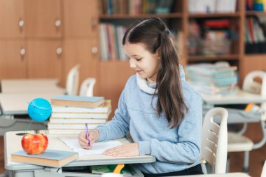 Öğrenci bir kız, renkli kitaplar, bir elma ve bir küre ile çevrili masasında öğrenme ve yaratıcılık sevincini somutlaştırarak mutlu bir şekilde çizim yapıyor..