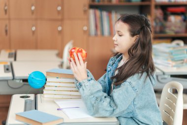 Bir liseli kız canlı bir sınıfta masasında oturuyor, elinde bir elmayla küreyi izliyor. Etrafı kitaplar ve okul malzemeleriyle çevrili olarak derslerine odaklanmış durumda..