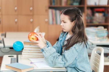 Öğrenme kaynaklarıyla dolu rahat bir sınıfta, bir öğrenci masasında otururken parlak kırmızı bir elmayı inceler, ilham verici bir eğitim ve keşif gününe hazırdır..