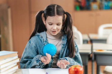 Parlak ışıklandırılmış sınıf canlanıyor ve liseli bir kız öğrenim materyallerini mutlulukla keşfediyor. Yaratıcılık dolu elleriyle, kitaplarla ve sanatla çevrili eğitim yolculuğunun tadını çıkarıyor..