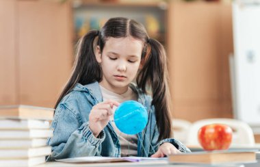 Odaklanmış bir liseli kız, öğrenmeye dalmış mavi bir küreyi inceliyor. Renkli elmalar ve kitap yığınlarıyla çevrili bir masada oturuyor. Heyecan verici bir okul gününe hazır..