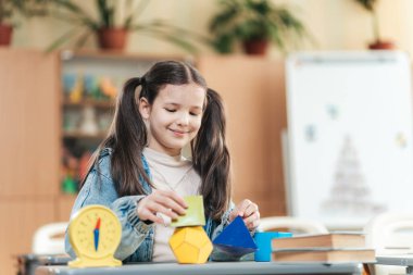 Canlı bir sınıfta, örgülü saçlı bir kız öğrenci renkli geometrik şekilleri düzenlerken gülümsüyor. Etrafı kitaplarla ve eğitim araçlarıyla çevrili olarak öğrenme sevincini kucaklıyor..