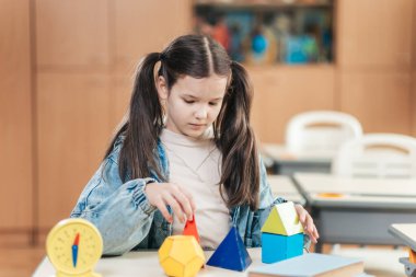 Saçları örgülü odaklanmış bir okul kızı sınıfında renkli geometrik şekiller düzenler. Lyceum 'da öğrenme aktivitesiyle yakından ilgilidir..