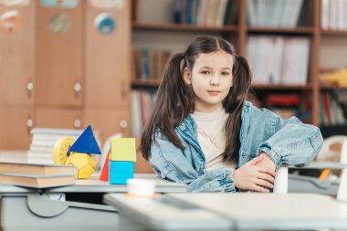 Bir liseli kız, eğitim malzemeleriyle dolu canlı bir sınıfta masasında güven içinde oturuyor. Düşünceli bir şekilde, sınıf arkadaşlarıyla ilham verici bir gün için hazır..