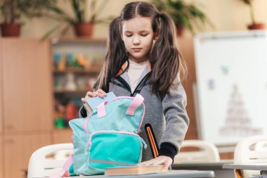 Canlı bir sınıfta, atkuyruklu bir kız öğrenci canlı sırt çantasını düzenlerken derslerine odaklanır. Öğrenme heyecanı havayı doldurur.