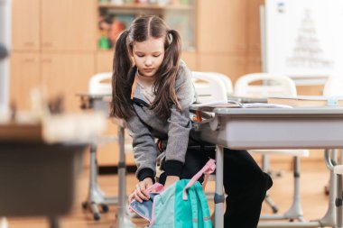 Canlı bir sınıfta, genç bir liseli kız renkli bir sırt çantasından malzemelerini toplamak için eğilir. Parlak masalar onu çevrelemiş, keşif ve arkadaşlık alanını sembolize ediyor..