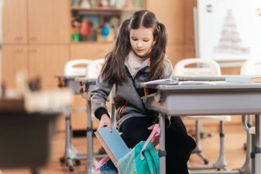 Genç bir öğrenci parlak bir sınıftaki bir masada oturur, derslerine odaklanırken kitapları dikkatlice sırt çantasına koyar. Atmosfer eğitim ve merakla dolu..