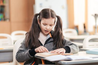 Genç bir öğrenci masasında oturmuş, derslerine odaklanmış. O, canlı sınıfında yeni bir bilgi keşfederken yüzünde bir merak bir bakışla, açık bir kitaptan okur..