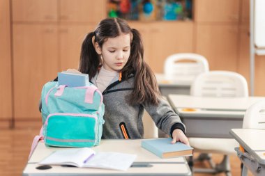 Odaklanmış bir öğrenci, parlak bir okul sınıfında sırt çantasını ayarlamak için kitaplarını masanın üzerine yerleştirir. Atmosfer öğrenme ve keşfetme enerjisiyle doludur..