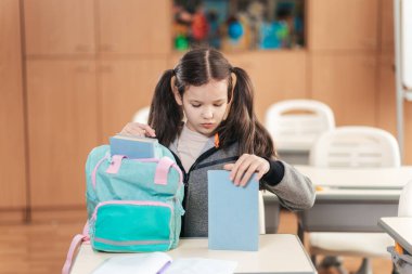 Genç bir liseli kız, iyi aydınlatılmış bir sınıfta masasında oturuyor, sırt çantasını dikkatlice açıyor. Okul malzemelerine odaklanmış, öğrenme gününü kucaklamaya hazır..