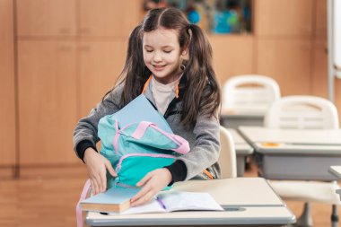 Canlı bir sınıf ortamında, örgülü saçları olan bir kız öğrenci, çalışmalarına dalmaya hazırlanırken masayı hevesle düzenliyor, kitap ve malzemelerini çıkarıyor..
