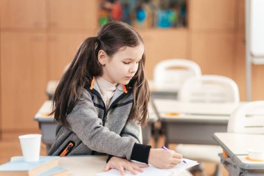 Öğrenmenin ve çocukluk keşiflerinin neşesini somutlaştıran, renkli süslemelerle dolu parlak bir sınıfla çevrili, masasına özenle notlar yazan bir kız öğrenci..