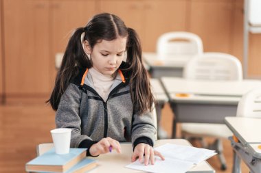 Genç bir liseli kız masasında oturmuş, yanında kitapları ve fincanı ile derslerine odaklanmış, hareketli bir sınıfta çocukluk öğreniminin özünü yakalıyor..