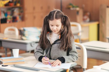 Genç öğrenci masasında oturuyor, özenle defterine yazıyor. Kitaplar ve renkli sınıf malzemeleriyle çevrelenmiş, merakı ve eğitim ruhunu somutlaştırıyor..