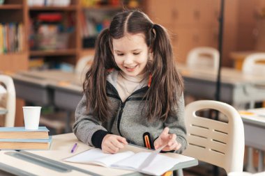 Uzun saçlı neşeli bir okul kızı masasında çalışırken gülümsüyor. Etrafı kitaplarla ve renkli kırtasiyelerle çevrili. Çocukluk öğrenme ruhunu somutlaştırıyor..