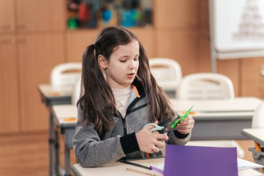 Öğrenme materyalleriyle dolu canlı bir sınıfta, genç bir kız, okul masasındaki zanaat projesine dalmış, kağıttan renkli şekiller yaratmak için dikkatle makas kullanıyor.