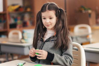 Genç bir öğrenci masasında oturuyor, elleriyle renkli bir şey yaratmaya odaklanmış. Gülümserken atkuyrukları sekiyor. Etrafı keşifleri teşvik eden hareketli bir sınıf atmosferiyle çevrili..