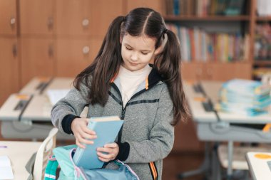 Canlı bir sınıfta, at kuyruklu genç bir öğrenci kitabını hevesle renkli bir sırt çantasına koyar. Masalar düzenli, sıcak bir öğrenme ortamı yaratıyorlar..