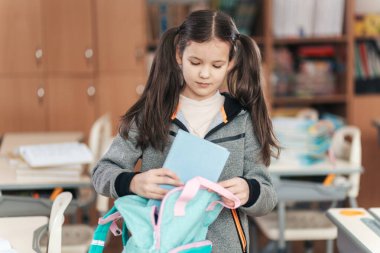 Atkuyruklu genç bir öğrenci okul malzemeleriyle dolu sırt çantasını dikkatlice düzenliyor ve canlı bir sınıf ortamında öğrenme gününe hazırlanıyor..
