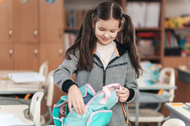 Sırt çantasını sınıf masasında düzenlerken saçları örgülü neşeli bir kız öğrenci gülümsüyor. Canlı ortam, okulun heyecanını yakalayan öğrenme materyalleriyle dolu..