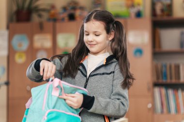 Bir liseli kız, heyecan ve merakla dolu sırt çantasını sınıfta mutlu bir şekilde düzenliyor. Parlak posterler duvarları süslüyor, canlı bir öğrenme ortamı yaratıyor..