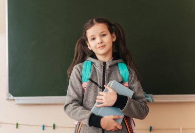 Genç öğrenci, okul gününe başlamak ve büyümek için sabırsızlanan yeşil bir kara tahtanın önünde dururken kitaplarını heyecanla kucaklıyor..