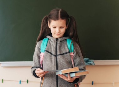 Liseli bir kız, neşeli bir sınıfta elinde kitaplarla, sırt çantasıyla duruyor ve öğrenme materyallerini inceliyor. Çalışmalarında yeni bir başlangıç için heyecanlı..