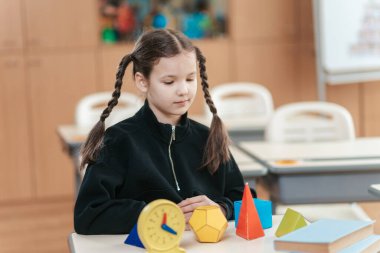 Düşünceli bir liseli kız masasında oturuyor, çeşitli geometrik şekillere odaklanmış, öğrenme kaynakları ve neşeli renklerle dolu canlı bir sınıfta kavramları keşfediyor..