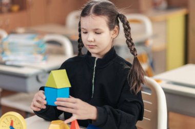 Odaklanmış bir liseli kız masasında canlı bir sınıfla çevrili renkli geometrik şekiller yığıyor. Çalışma sırasında azim ve yaratıcılık sergiliyor..