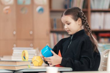 Örgülü saçlı bir kız öğrenci, öğrenme aşkını vurgulayarak, masasında canlı bir sınıf atmosferinde kitaplarla otururken geometrik şekilleri keşfetmeye odaklanır..