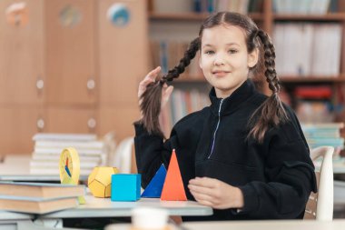 Parlak gözlü liseli kız çalışmalarına renkli geometrik şekillerle dolu bir masada dalmış, canlı bir sınıf ortamında öğrenme hevesini sergiliyor..