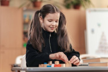 Genç bir liseli kız masasında oturuyor, renkli yapı taşlarını yığmaktan mutlu. Odaklanmış ve gülümsüyor. Eğitici materyallerle dolu parlak sınıfında öğrenme deneyiminin tadını çıkarıyor..