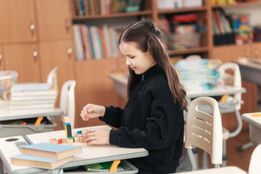 Canlı bir sınıfta, uzun saçlı bir kız öğrenci renkli blokları olan bir bina inşa etmeye odaklanır..