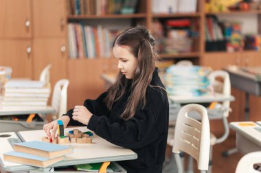 Genç bir kız masasında renkli bloklarla çeşitli şekiller oluşturmaya konsantre olur. Canlı sınıf, öğrenme ve keşfin özünü yakalayan kitaplar ve malzemelerle dolu..