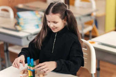 Okullu bir kız sınıf arkadaşları tarafından çevrelenmiş renkli ahşap yapı taşları kullanarak canlı bir yapı inşa etmeye odaklanırken, parlak ve neşeli sınıflar kahkahalarla dolu..