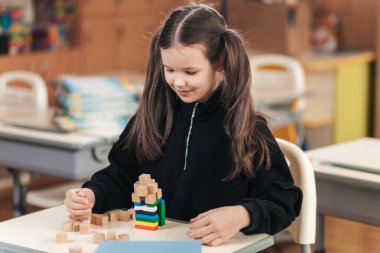 Canlı bir sınıfta, örgülü saçlı bir kız öğrenci neşeyle ahşap bloklardan bir kule inşa ediyor. Kibar ifadesi, eğlenceli bir öğrenme sürecinde parlıyor..