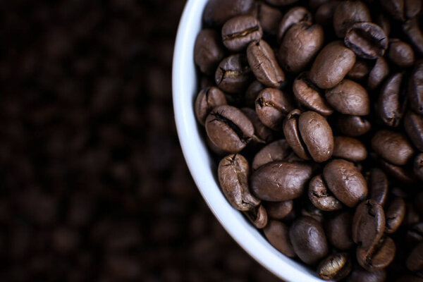White cup full of coffee beans close up against the coffee beans background. Coffee mug. Morning espresso.