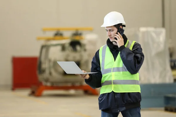 Portrait of a man , factory engineer in work clothes holding laptop and phone, controlling the work process at the helicopter manufacturer.