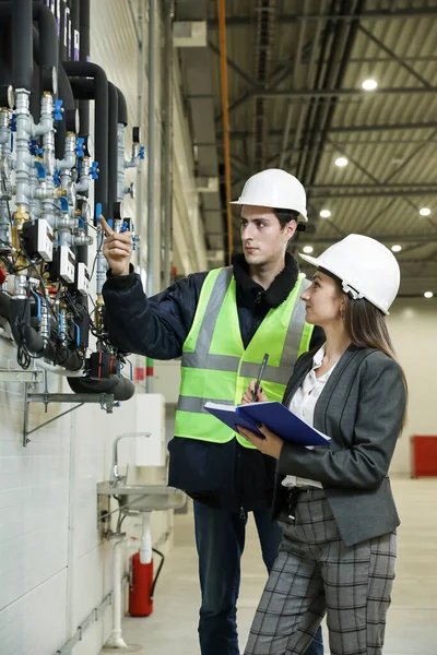 Portrait of a female factory manager in a white hard hat and business suit and factory engineer in work clothes. Controlling the work process in the manufacture.