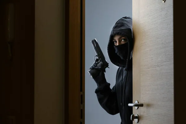 Thief broke into the apartment. House robbery by woman in a black jacket and black mask black gun and crowbar. Burglar in a mask. Thief in a mask trying to break into other people\'s house.